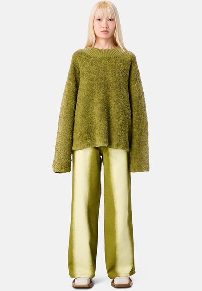 Vihreä teksturoitu neulepaita pitkillä hihoilla yhdistettynä vaalean vihreisiin satiinisiin leveälahkeisiin housuihin. Malli seisoo yksinkertaista taustaa vasten.