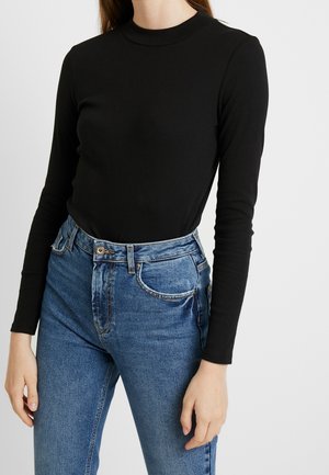 Person iført en tætsiddende sort langærmet top, der er stukket ned i højttaljede blå denimjeans med frontlommer og knaplukning.