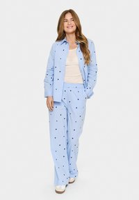 Set di pigiama azzurro chiaro composto da una camicia con bottoni e pantaloni con cintura elastica, decorato con piccoli cuori blu navy, abbinato a scarpe bianche slip-on.