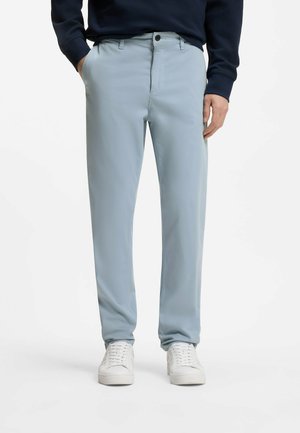 Homme portant un pantalon bleu clair à coupe droite, un sweat-shirt bleu marine et des baskets blanches, debout avec une main dans la poche sur fond blanc.