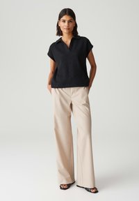 Schwarzes kurzärmeliges Poloshirt mit strukturiertem Design und V-Ausschnitt. Kombiniert mit beigen weit geschnittenen Hosen und schwarzen Riemchensandalen.