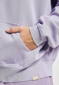 Sweat à capuche violet clair avec une poche avant, des poignets et un ourlet côtelés, arborant une petite étiquette marquée "DEF" sur le côté inférieur. Texture lisse.