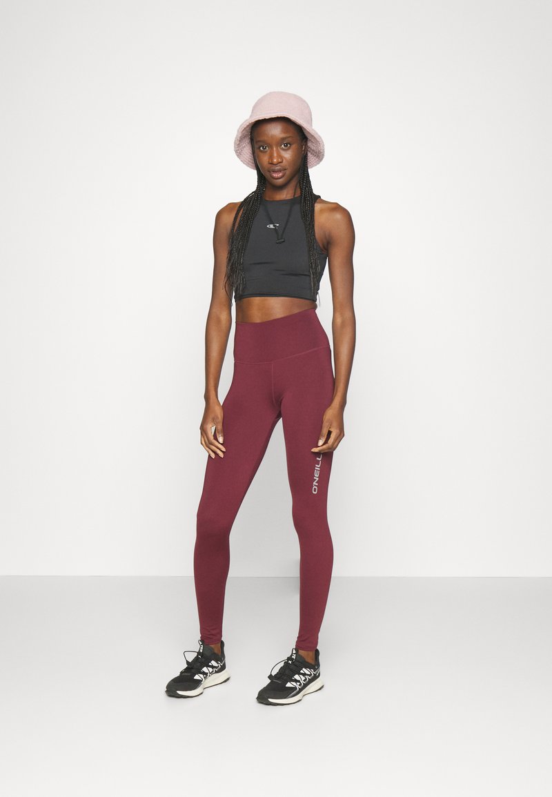 Schwarzes Croptop, kastanienbraune High-Waist-Leggings mit Logodetail, schwarze Sportschuhe und ein rosa Bucket Hat. Glatte Stofftextur.