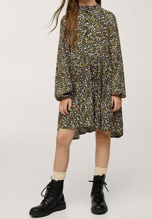 Robe noire à fleurs avec des manches longues, présentant des motifs jaunes et blancs. Accompagnée de bottes noires à lacets et de chaussettes crème jusqu'à la cheville.
