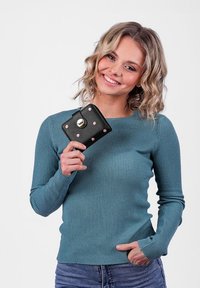 Black wallet with pink stud accents and gold hardware held by a person wearing a ribbed teal top. Background is plain white.