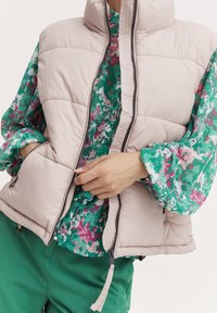 Vest puf roz deschis cu guler înalt; are un zipper și buzunare laterale. Sub vest, o bluză florală multicoloră cu bază verde și accente roz.