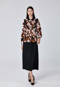 Blouse florale marron avec un col noir à volants, associée à une jupe plissée noire et des chaussures à talons hauts noires. Fond simple.