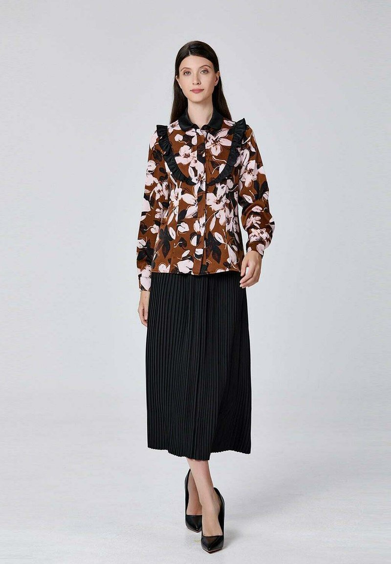 Blouse florale marron avec un col noir à volants, associée à une jupe plissée noire et des chaussures à talons hauts noires. Fond simple.