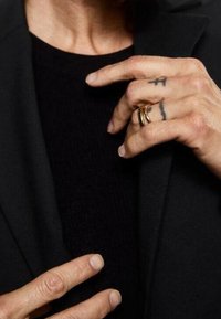 Anneaux en métal doré empilés sur une main tatouée, reposant contre un blazer noir et une chemise noire texturée. Teint clair.