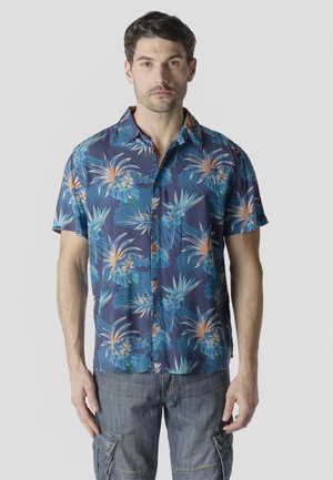 Hombre con camisa azul de manga corta abotonada con estampado tropical floral en naranja y azul, y pantalones cargo grises, de pie contra un fondo liso.