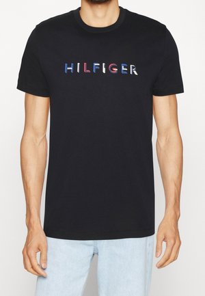 Mann trägt ein schwarzes T-Shirt mit kurzen Ärmeln mit dem Wort "HILFIGER" in roten, weißen und blauen Buchstaben auf der Brust, kombiniert mit hellblauen Jeans.