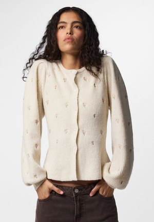 Femme aux longs cheveux bouclés portant un cardigan en maille crème avec de petites broderies florales et un pantalon foncé, mains dans les poches, expression neutre.
