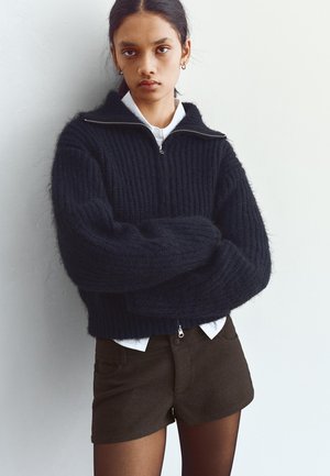 Črni pleteni pulover z visokim ovratnikom, zadrgo in rebrasto teksturo. Kombiniran z rjavimi kratkimi hlačami in belo srajco pod njim.