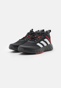 Baskets de sport noires avec un dessus en maille et synthétique, présentant trois rayures blanches, des accents rouges et une semelle en caoutchouc noir texturé.