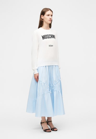 Γυναίκα που φοράει λευκό φούτερ Moschino Milano, φωτεινή μπλε midi φούστα με στρώσεις και μαύρα σανδάλια με τακούνι, στέκεται μπροστά σε λευκό φόντο.