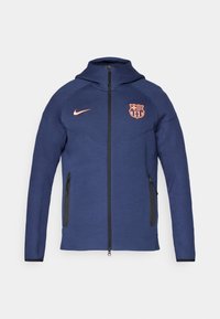 Veste à zip intégral bleu marine avec capuche, deux poches latérales zippées, logo Nike orange et blason du club FC Barcelona sur le devant.