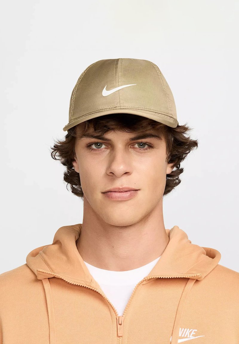 Cappello khaki con un design strutturato, caratterizzato da un logo Nike swoosh bianco sulla parte anteriore. Uomo con capelli ricci, che indossa una felpa con zip di colore pesca sotto.