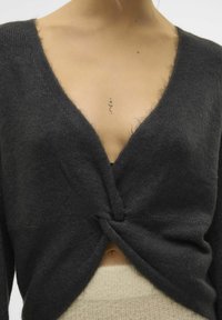 Cardigan en tricot noir avec un décolleté en V profond et un design à l'avant torsadé, présentant une texture douce et une légère peluchabilité. Superposé sur un top beige.