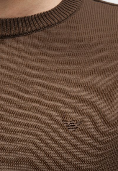 Lähivõte pruunist kampsunist, millel on soonikkoes ümarselg ja väike tikitud logo rinnal.