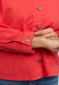 Korallfärgad jacka med silverknappar, krage och en bröstficka. Tyget ser lätt och mjukt ut, med detaljer vid ärmsluten.