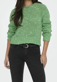 Zielony, dziergany sweter o luźnym kroju, zdobiony żółto-białymi cętkami, zestawiony z czarnymi, wysokim stanem spodniami jeansowymi i czarnym paskiem.
