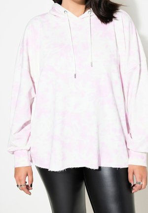 Person iført en løs lyserød og hvid tie-dye hættetrøje med snore og sorte læderbukser, stående mod en ensfarvet baggrund.
