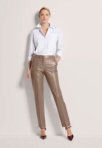 Vrouw die tegen de muur staat in een witte blouse met knopen, taupe leren broek en donkere puntige hakken met enkelbandjes.