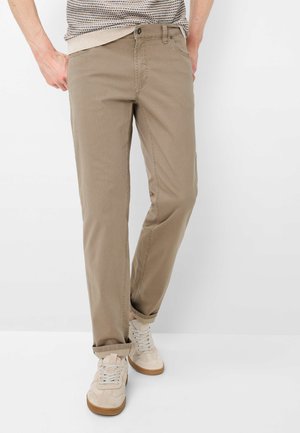 Mann trägt beige gerade geschnittene Hose mit aufgerollten Bündchen und weißen Sneakers vor weißem Hintergrund.