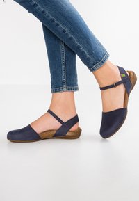 Granatowe buty wsuwane z regulowanym paskiem w okolicy kostki, z teksturowaną brązową korkową podeszwą i zaokrąglonym czubkiem.