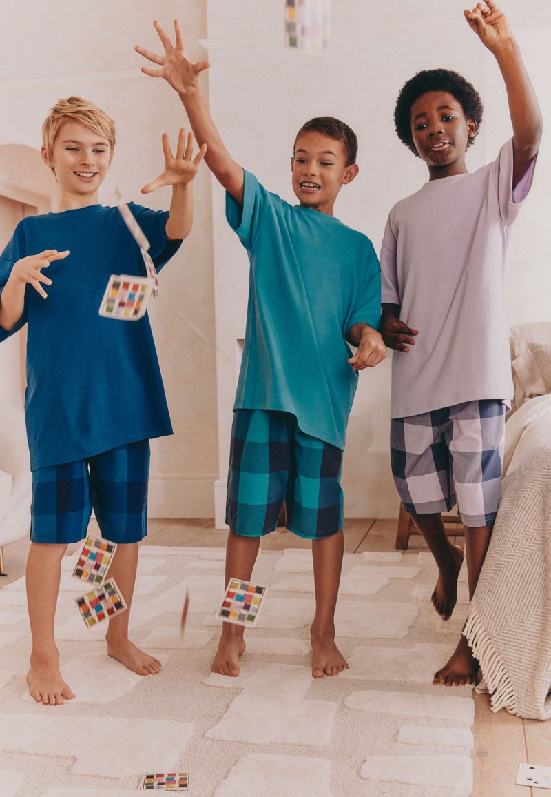 Tre drenge iført pyjamas kaster begejstret spillekort inde i et hyggeligt rum med et lyst tæppe og neutrale vægge.