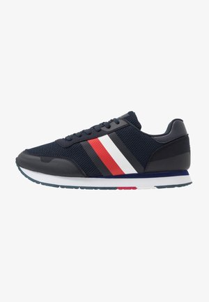 Scarpa da ginnastica blu navy con tomaia in mesh, sovrapposizioni nere e strisce rosse, bianche e nere sul lato, suola bianca con accenti blu e rossi.