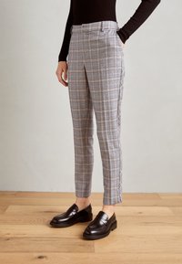 Kostkované kalhoty v šedé barvě s růžovými akcenty, střih na míru, rovné nohavice, postranní kapsy, nošené s černými mokasínami. Dřevěná podlaha v pozadí.