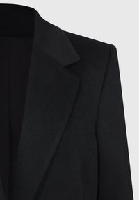 Blazer negro con un cuello estructurado, hecho de tejido texturizado. Presenta solapas afiladas y una silueta limpia y entallada.