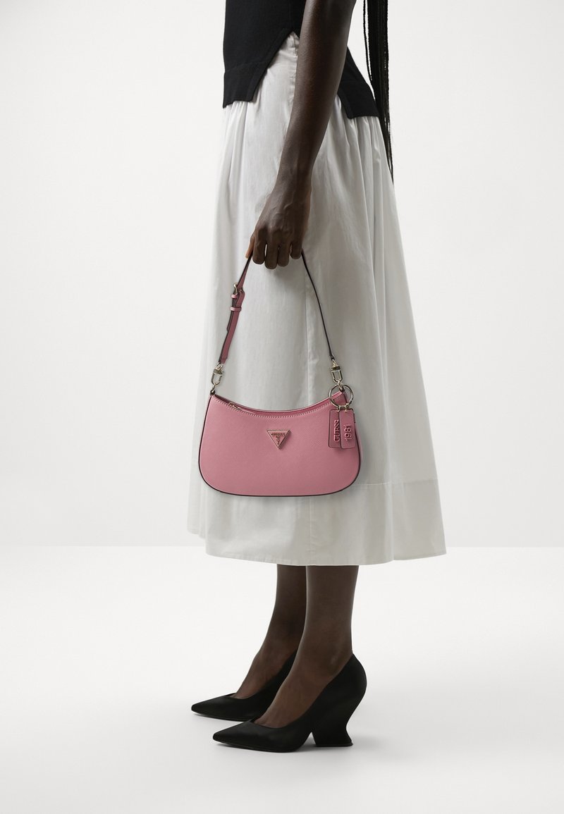 Femme en haut noir, jupe blanche et talons noirs tenant un petit sac à main rose avec des étiquettes sur un fond blanc uni.