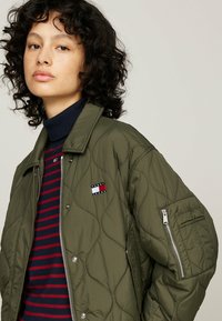 Chaqueta acolchada de color verde olivo con un bolsillo con cremallera y un logo bordado, combinada con un cuello de tortuga a rayas en tonos azul marino y rojo debajo.