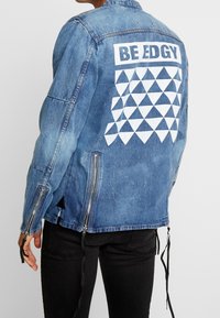 Person som bär en blå denimjacka med texten "BE EDGY" och vita trianglar på baksidan, tillsammans med svarta byxor, står mot en enfärgad bakgrund.