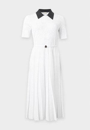 Frau mit langen schwarzen Haaren, die ein weißes kurzärmeliges Kleid mit dunklem Kragen und beigen Sandalen trägt, steht mit einer Hand in der Hüfte.