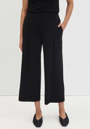 Donna che indossa pantaloni neri a gamba larga corti con tasche laterali e ballerine nere con punta su sfondo chiaro e uniformemente colorato.