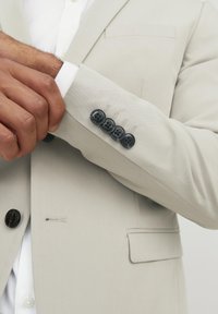 Hellgrauer Blazer mit glatter Textur, ausgestattet mit schwarzen Knöpfen an den Manschetten und einer einzelnen Brusttasche. Sichtbares Knopfloch-Detail.