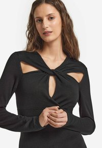 Svart långärmad topp med utskärningsdetaljer vid axlarna och en knutdesign vid halsringningen, tillverkad av mjukt, texturerat tyg.