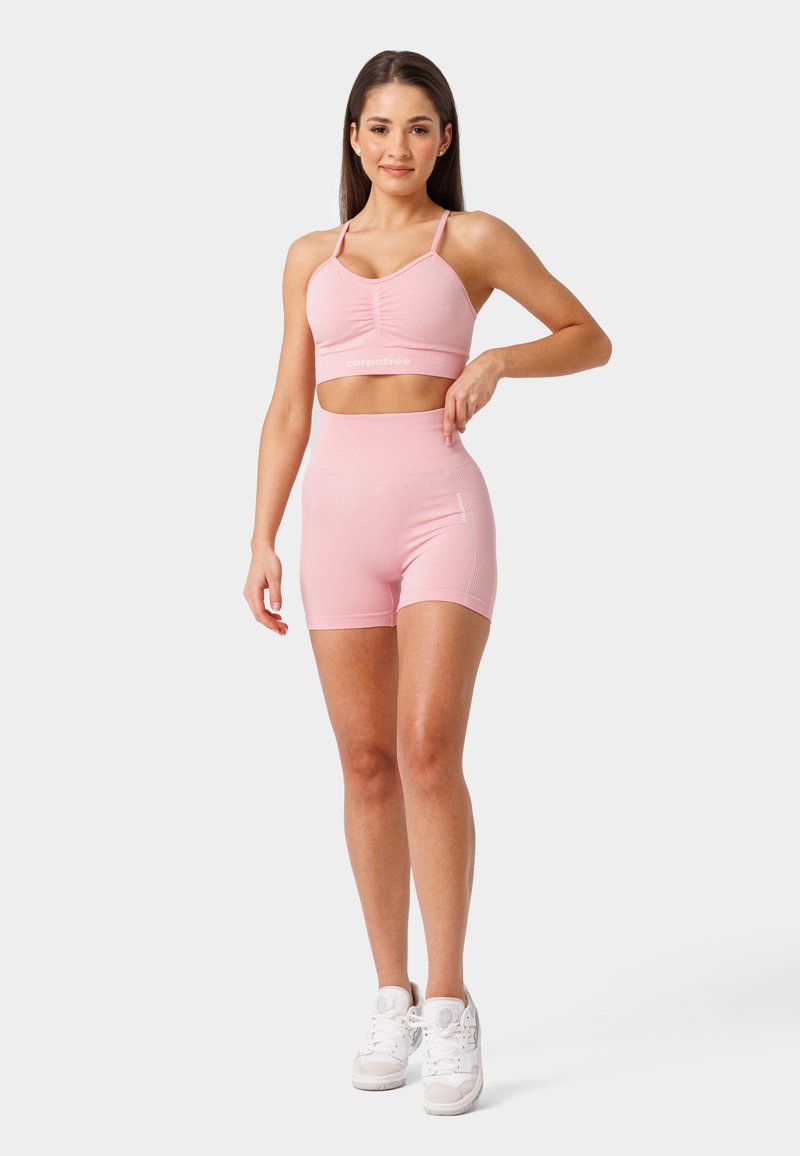 Sujetador deportivo rosa claro con un diseño fruncido y logo, combinado con pantalones cortos ajustados de cintura alta. Los materiales son elásticos y suaves.
