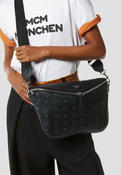 Persoon draagt een wit MCM München T-shirt en zwarte broek en houdt een zwarte MCM crossbodytas met zilveren ritssluiting vast.