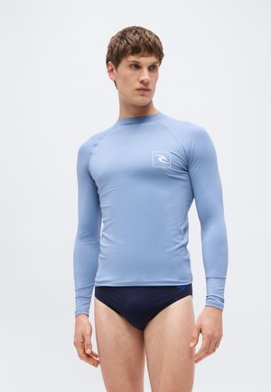 Junger Mann, der ein enganliegendes hellblaues langärmeliges Schwimmshirt und dunkelblaue Badehosen trägt, steht vor einem schlichten Hintergrund.