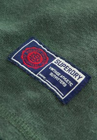 Žalias tekstūruotas audinys su mėlyna ir balta stačiakampio formos etikete. Etiketeje yra užrašai "SUPERDRY", "VINTAGE ATHLETIC OUTFITTERS" ir raudonas apvalus emblema.