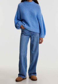 Personne portant un pull en tricot bleu, un jean large bleu et des chaussures brunes à enfiler, se tenant avec une main dans la poche.