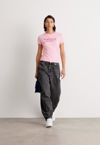 Pink cotton T-shirt with "tommy jeans" logo, paired with loose-fitting black denim pants and white sneakers. Model carries a blue bag.