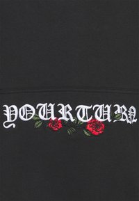 Felpa nera con testo ricamato bianco che recita "IL TUO TURNO," decorata con rose rosse e foglie verdi.