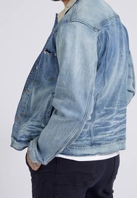 Light blue denim jacket with distressed detailing, front pockets, and button closures. Visible stitching and a relaxed fit.