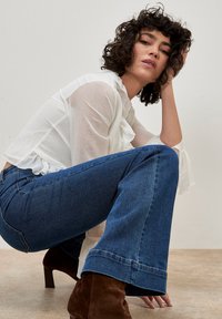 Blusa bianca e testurizzata con ruffles, abbinata a jeans a zampa d'elefante blu. La modella indossa stivaletti alla caviglia in camoscio marrone. Sfondo semplice e neutro.