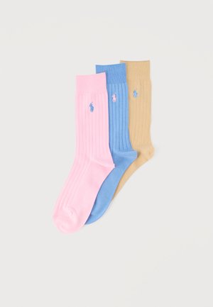 Trois chaussettes côtelées en rose, bleu et beige, chacune avec un petit logo brodé près du haut, posées à plat sur un fond blanc.
